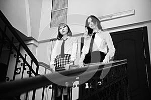 Two sad schoolgirls dressed in white shirts and black ties are standing on a school staircase near the railing and looking down.