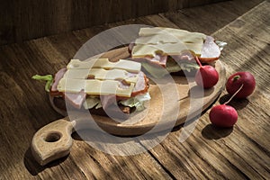 two sandwiches with cheese, ham and lettuce on a cutting board on a wooden background lit by natural morning light from the window