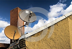 Two satellite dishes in front of a red brick chimney on the sloping roof of a house