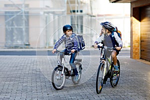 Two school kid boys in safety helmet riding with bike in the city with backpacks. Happy children in colorful clothes