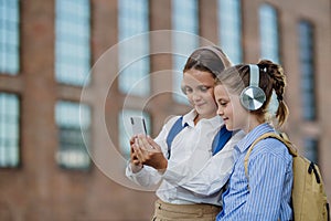 Two schoolgirls checking social media on smartphone in front of school. Friends with headphones are watching a video.