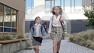 Two schoolgirls of different ages with backpacks go to school, holding hands. The older sister takes the younger one to