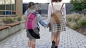Two schoolgirls of different ages with backpacks go to school, holding hands. The older sister takes the younger one to