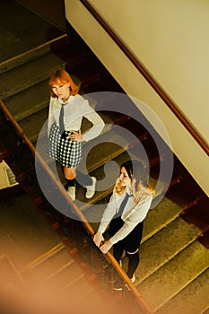 Two schoolgirls, dressed in their school uniforms, standing near the staircase railing. They are friends