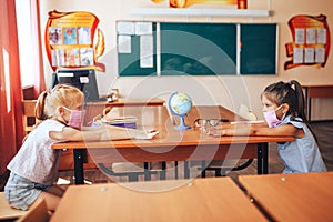 Two schoolgirls in medical masks are sitting at a school desk, opposite each other, group session, back to school, teaching