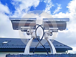 Two security cameras on the fence against the roof of the house and the blue sky with clouds.