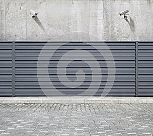 Two security cameras hanging on the concrete wall of the building, aimed at the space behind the fence