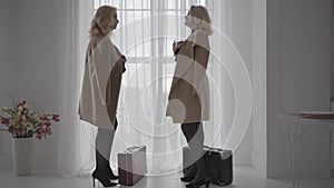 Two senior blonde Caucasian women in elegant beige coats walking with travel bags to the window. Rich senior