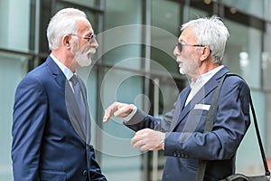 Two senior businessmen with gray hair and beard meeting and discussing in front of an office building. View from the side. Active