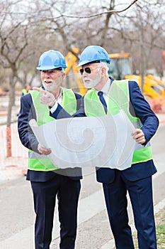 Two senior engineers or businessmen visiting construction site, looking at blueprints and discussing