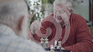 Two senior men playing chess sitting at home. Plump man in red shirt making a move. Caucasian old men neighbors friends