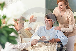 Two senior pensioners enjoying their leisure time together inside a private nursing home. Tender caretaker in uniform standing ne