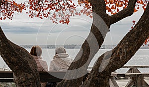 Two senior sat on a bench. Elderly femaile freinds in a autumnal park by the river