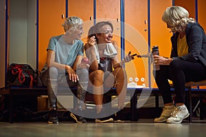 Two senior women with younger woman trainer sitting in gym dressing room having a conversation before workout