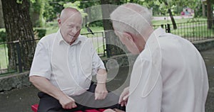 Two seniors friends playing game which hand is chess piece in on bench in summer