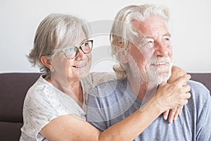 Two seniors and mature couple of people sitting on the sofa at home hugged looking at the camera happy with love and affection