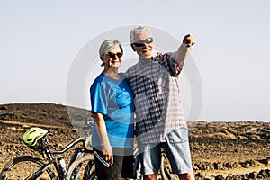 Two seniors together in mountain standing with their bikes at the background looking at something and man indicating something