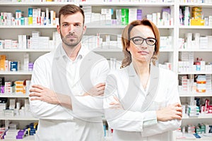 Two serious female and male pharmacists standing side by side looking directly at camera with arms crossed