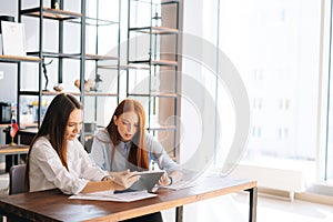 Two serious focused young business women working using digital tablet during meeting at desk with job documents at