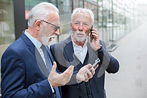 Two serious senior businessmen using mobile phones in front of an office building