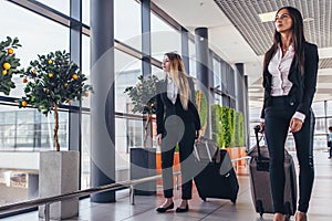 Two serious young female colleagues walking through airport passageway carrying suitcases