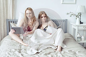 Two blonde girls in pyjamas having fun in the bedroom. Young females lying in bed using tablet.