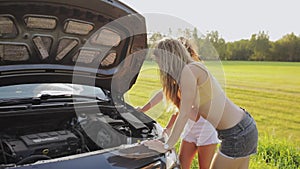 Two sexy girls in shorts and a t-shirt stand at the open hood of the car on the road in the woods, the field and try to