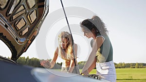 Two sexy girls in shorts and a t-shirt on sun stand at the open hood of the car on the road in the woods, the field. the