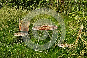 Two shabby and weathered rusty chairs and a table of metal in a wild garden with long green grass