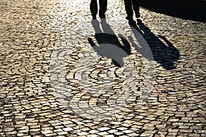 Shadows of two walking people projected on cobblestone pavement. Sunny summer morning.