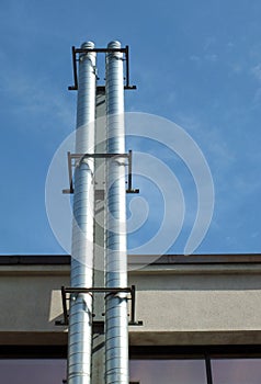 Two shiny steel metal industrial extraction chimneys on the side of a building against a blue summer sky with sunlight and shadow
