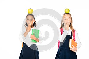 two shocked multicultural schoolgirls with apples on heads covering mouths with hands isolated on white.