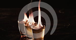 Two shot glasses with a clear liquid are ablaze with flames, set against a dark background