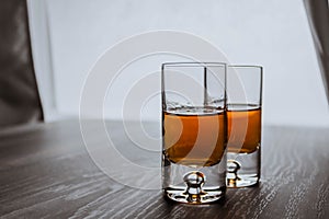 Two shot glasses with whiskey on a wooden background table next to window. Space for text