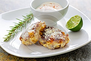 Two shrimp cakes on a white plate with a lemon garlic dipping sauces lemon wedge and a stem or rosemary on a wooden table.