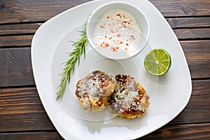 Two shrimp cakes on a white plate with a lemon garlic dipping sauces lemon wedge and a stem or rosemary on a wooden table.
