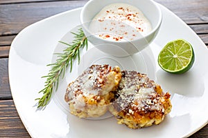 Two shrimp cakes on a white plate with a lemon garlic dipping sauces lemon wedge and a stem or rosemary on a wooden table.