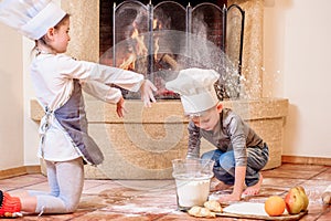 Two siblings - boy and girl - in chef`s hats near the fireplace sitting on the kitchen floor soiled with flour, playing with food
