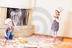 Two siblings - boy and girl - in chef`s hats near the fireplace sitting on the kitchen floor soiled with flour, playing with food