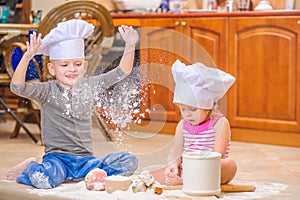 Two siblings - boy and girl - in chef`s hats sitting on the kitchen floor soiled with flour, playing with food, making mess and ha