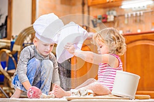 Two siblings - boy and girl - in chef`s hats sitting on the kitchen floor soiled with flour, playing with food, making mess and ha