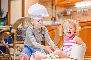 Two siblings - boy and girl - in chef`s hats sitting on the kitchen floor soiled with flour, playing with food, making mess and ha