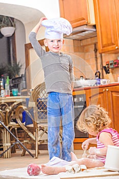 Two siblings - boy and girl - in chef`s hats sitting on the kitchen floor soiled with flour, playing with food, making mess and ha