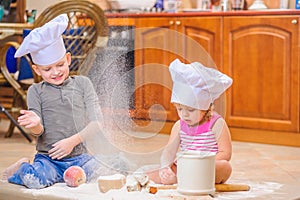 Two siblings - boy and girl - in chef`s hats sitting on the kitchen floor soiled with flour, playing with food, making mess and ha