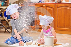 Two siblings - boy and girl - in chef`s hats sitting on the kitchen floor soiled with flour, playing with food, making mess and ha