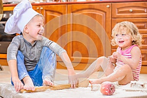 Two siblings - boy and girl - in chef`s hats sitting on the kitchen floor soiled with flour, playing with food, making mess and ha