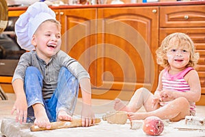 Two siblings - boy and girl - in chef`s hats sitting on the kitchen floor soiled with flour, playing with food, making mess and ha