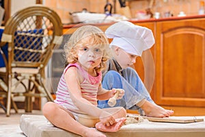 Two siblings - boy and girl - in chef`s hats sitting on the kitchen floor soiled with flour, playing with food, making mess and ha