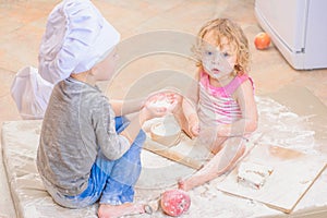 Two siblings - boy and girl - in chef`s hats sitting on the kitchen floor soiled with flour, playing with food, making mess and ha photo