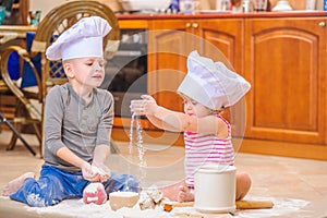 Two siblings - boy and girl - in chef`s hats sitting on the kitchen floor soiled with flour, playing with food, making mess and ha photo